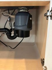 A gray and black garbage disposal unit installed under a kitchen sink with attached black plumbing pipes.