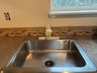 A stainless steel kitchen sink with a white faucet and a tiled backsplash, set against a plain wall with an exposed outlet.