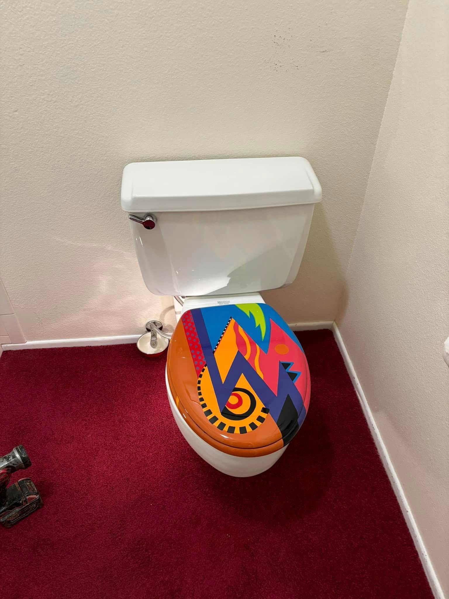 A toilet with a colorful, abstract patterned lid sitting on a deep red carpet.