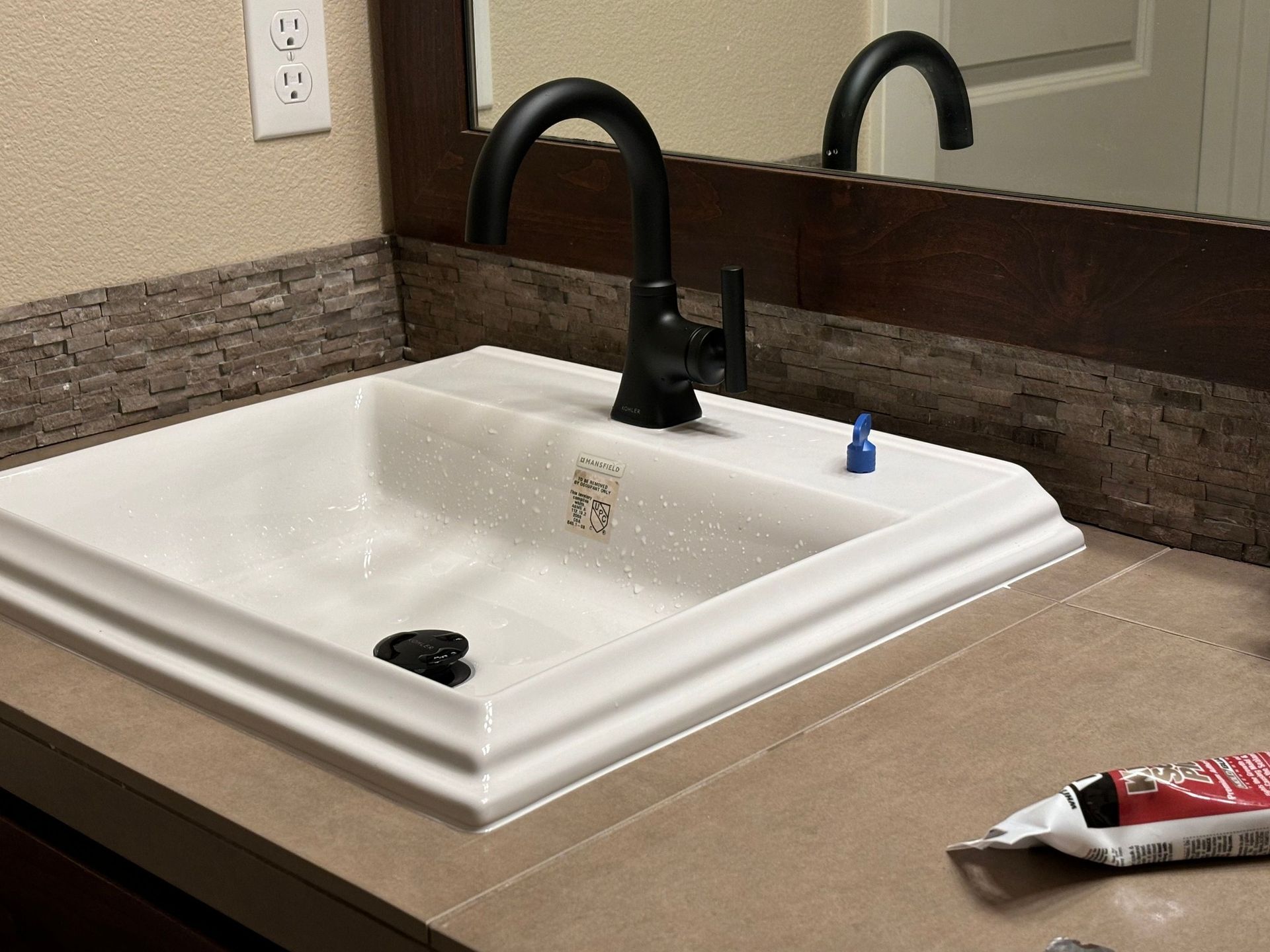 A white bathroom sink with a matte black faucet, set against a dark-tiled backsplash, with toothpaste on the counter.