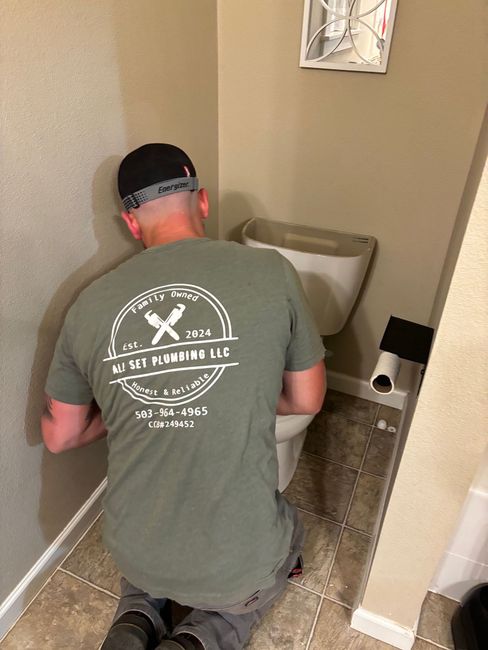 A person in an olive green t-shirt kneeling on a bathroom floor, inspecting or repairing a toilet.