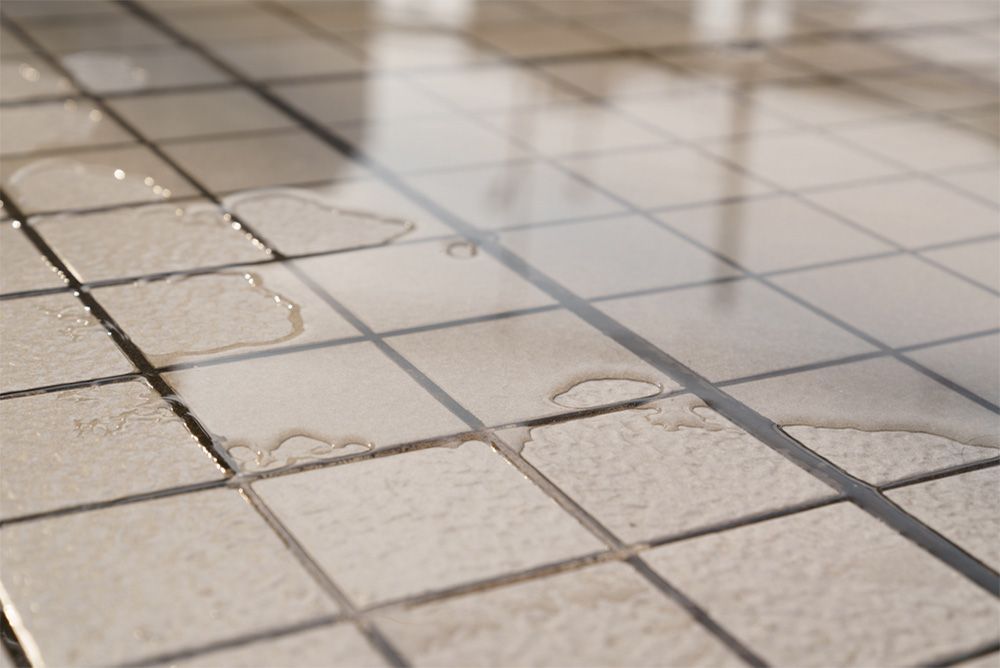 Water puddles scattered across light-colored, square bathroom or kitchen floor tiles.