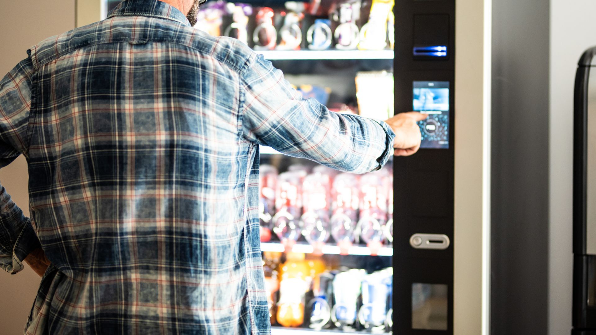 Person in plaid shirt using a vending machine's touchscreen, selecting a snack.