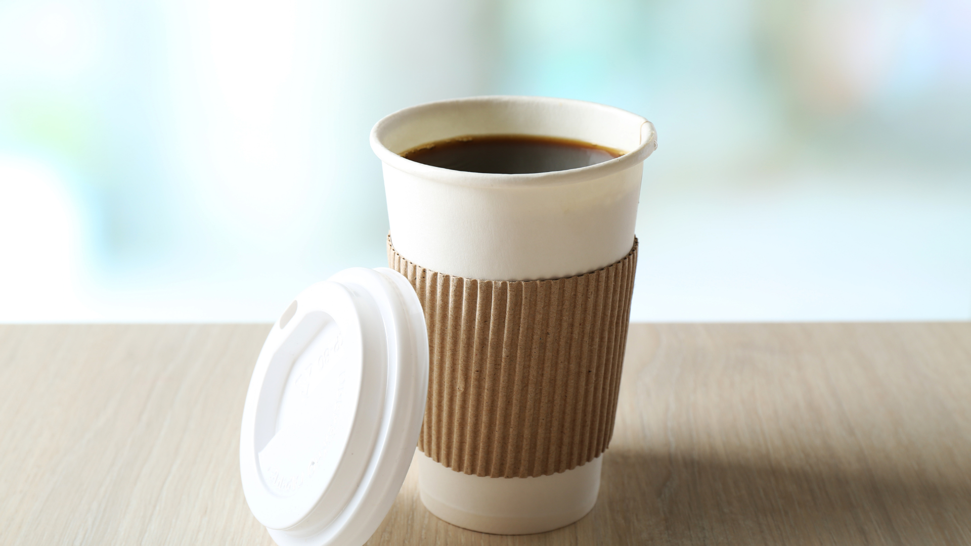 Coffee in a white paper cup with a brown sleeve, lid beside it, on a light wood surface.