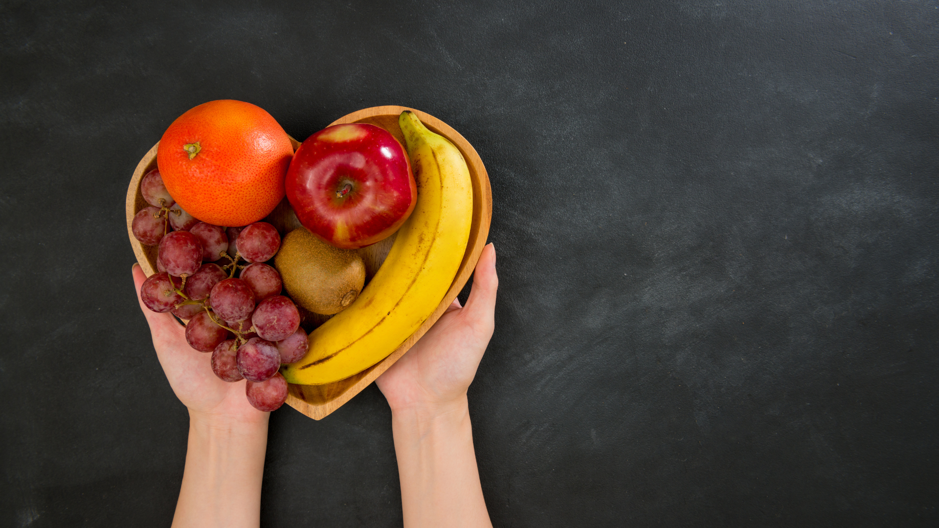 Hands holding a heart-shaped bowl of fruit: grapes, orange, apple, kiwi, and banana against a black background.