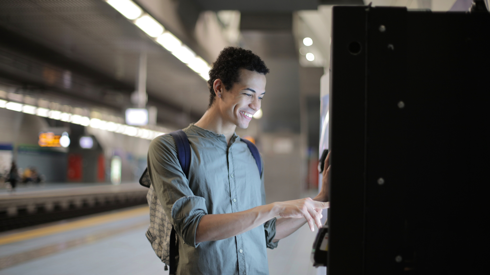 Smiling man at a subway kiosk, selecting options. He wears a backpack, standing on a platform.