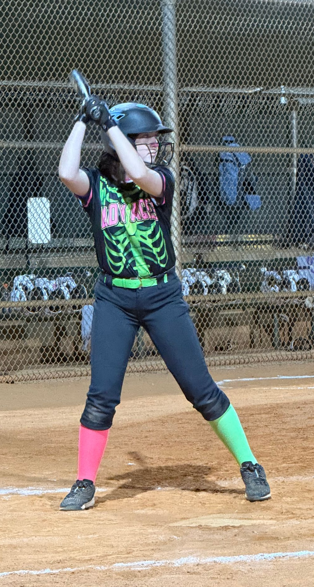 Softball batter in black helmet and colorful uniform standing at home plate, bat raised in a batting cage.