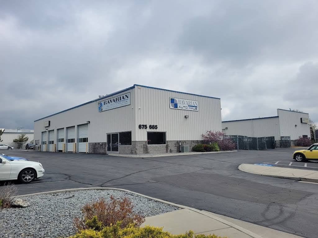 White industrial building with blue trim and signage, driveway, cars, and overcast sky. | Bavarian Auto Haus