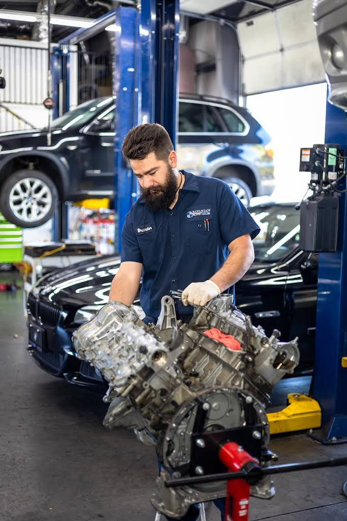 Mechanic holding an engine in a garage; a car is lifted, other vehicles present. | Bavarian Auto Haus