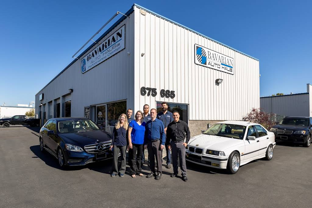 People standing in front of a white auto repair shop, with cars parked out front, on a sunny day. | Bavarian Auto Haus