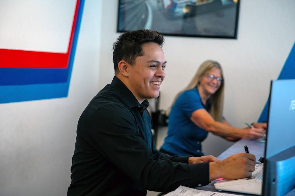 Man smiles, writing at a desk, with a woman in the background. Blue, white, and red decor. | Bavarian Auto Haus