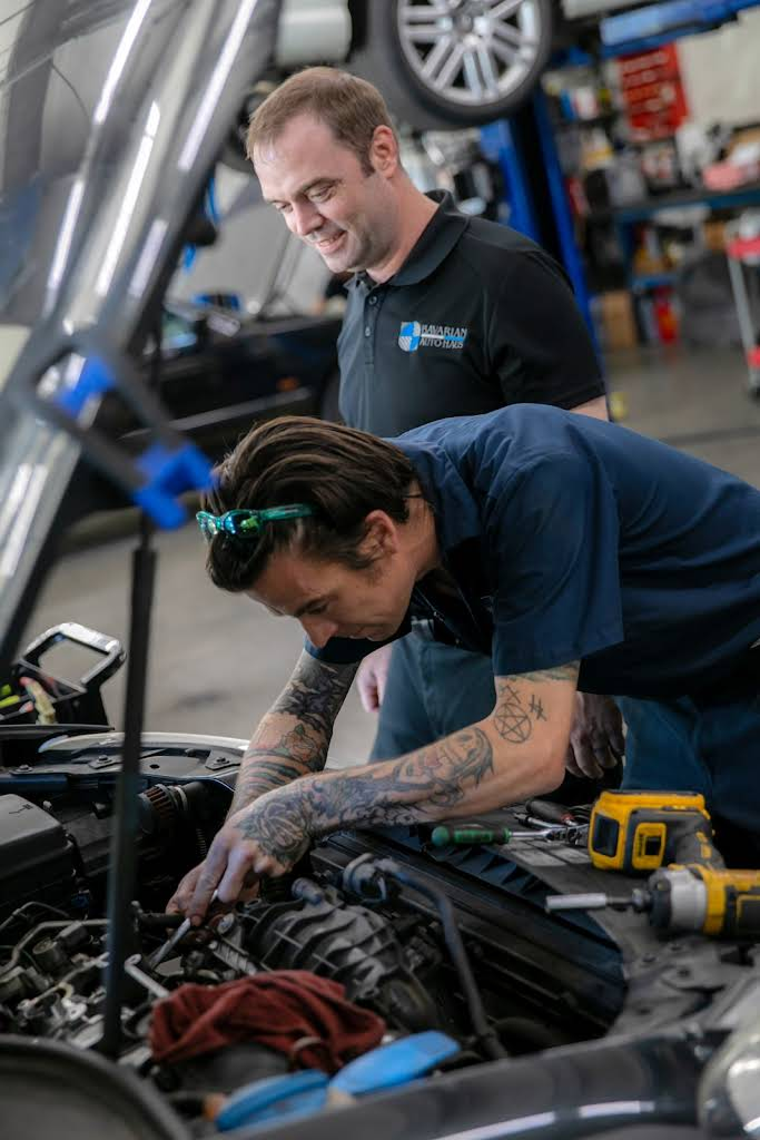 Two mechanics working on a car engine in a shop. One points at engine, the other watches. | Bavarian Auto Haus