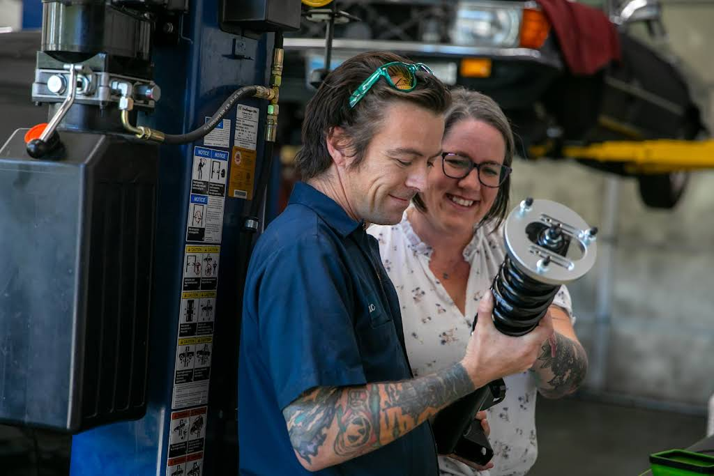 Mechanic and customer examining car suspension part in a garage, both smiling. | Bavarian Auto Haus