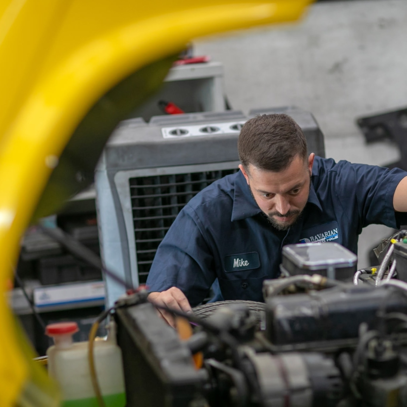 Mechanic working on car engine, looking intently. Yellow car hood frame, blue shirt, shop interior. | Bavarian Auto Haus