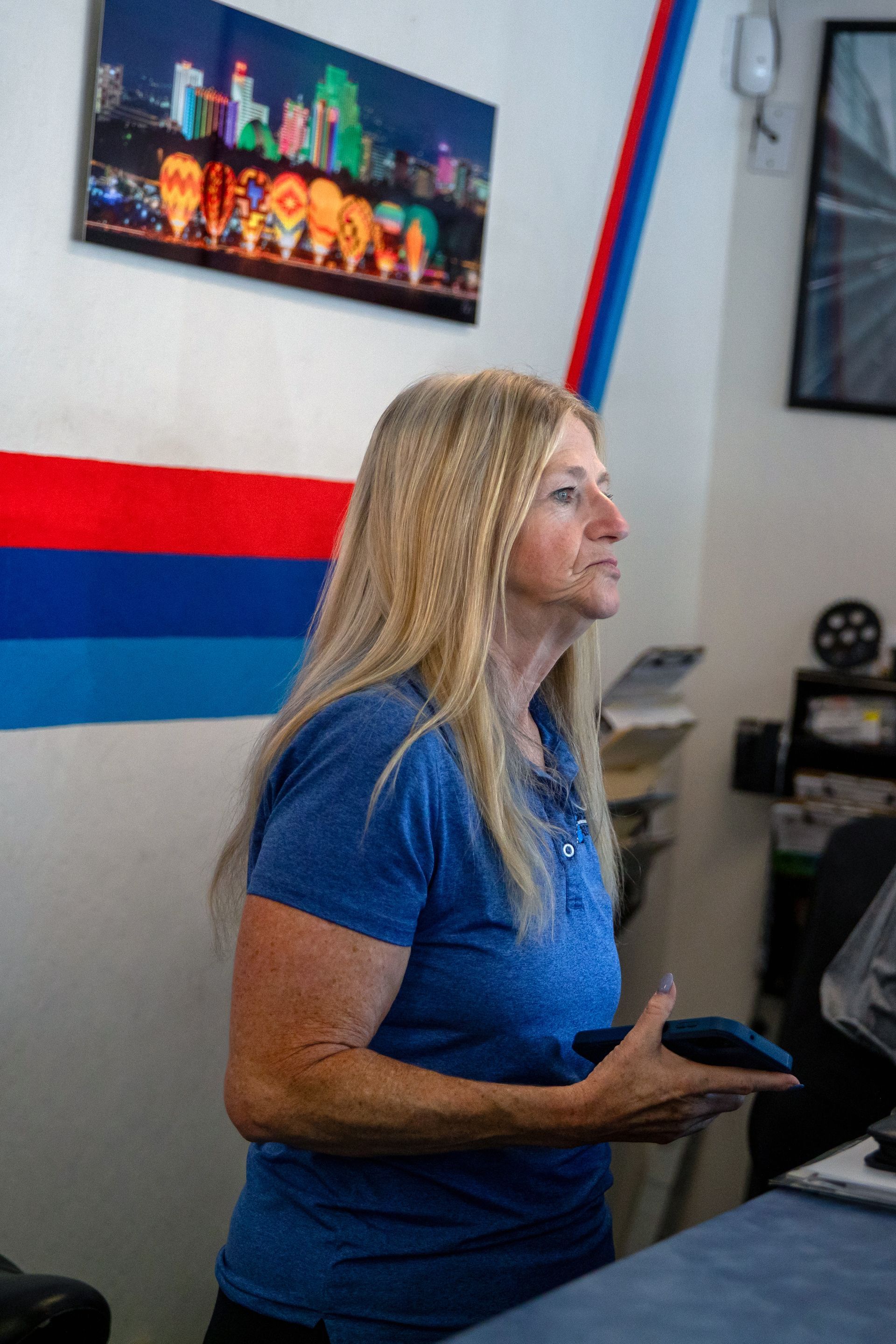A person with long blonde hair in a blue polo shirt holds a dark object while looking toward the right in an office. | Bavarian Auto Haus