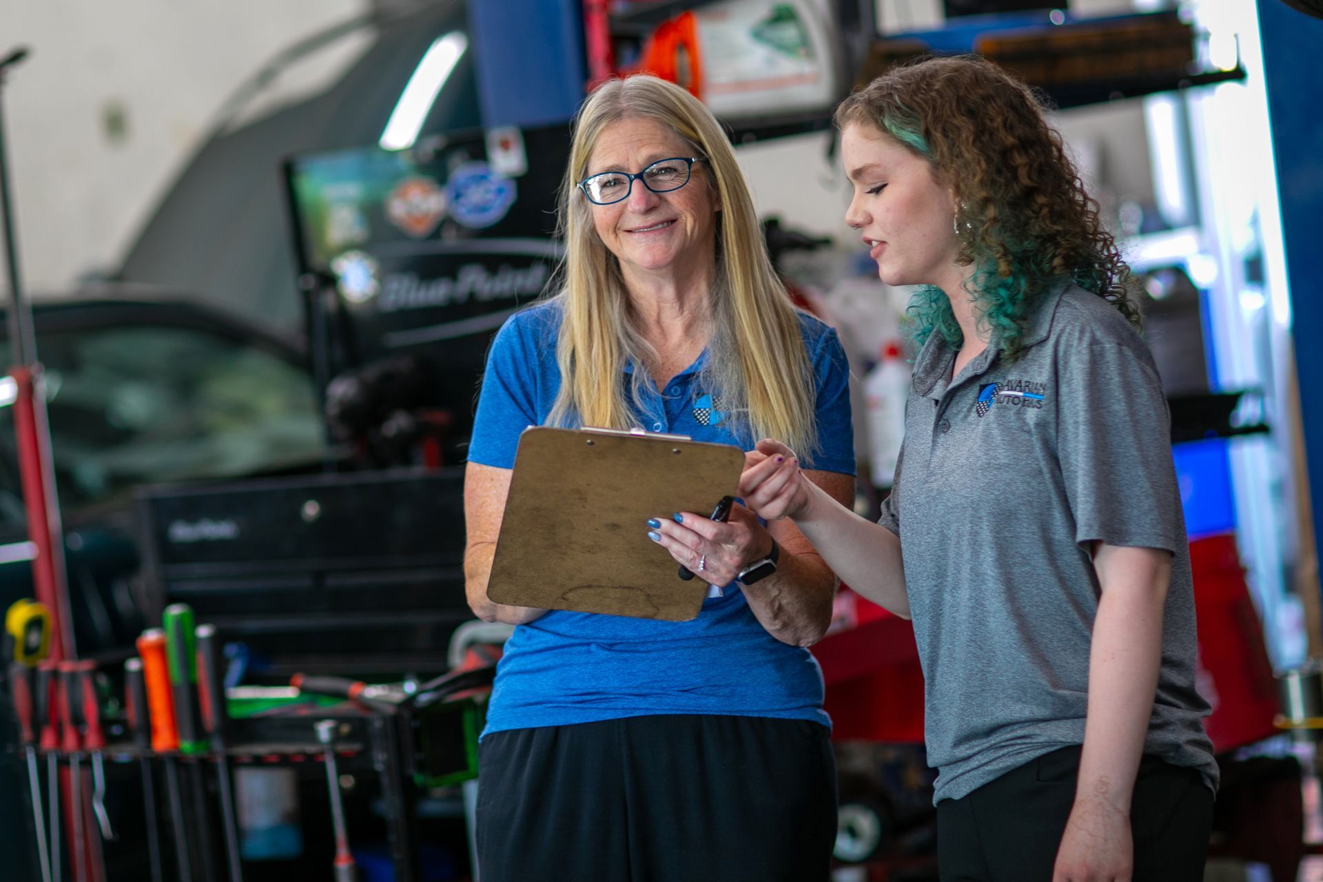 Two people in a garage workshop hold a clipboard and discuss technical work, wearing matching company shirts. | Bavarian Auto Haus