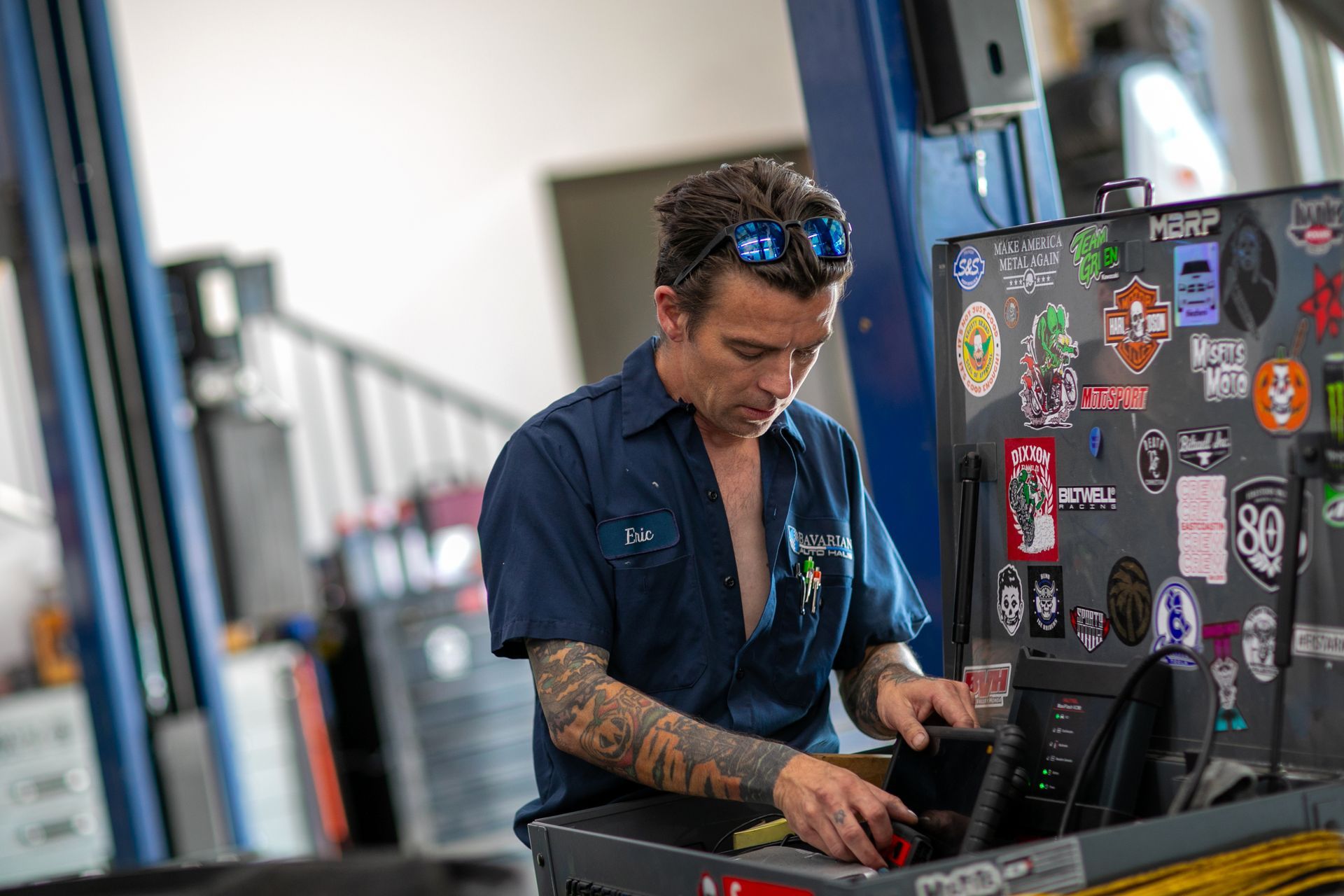A mechanic with arm tattoos works at a sticker-covered toolbox in a repair shop. | Bavarian Auto Haus