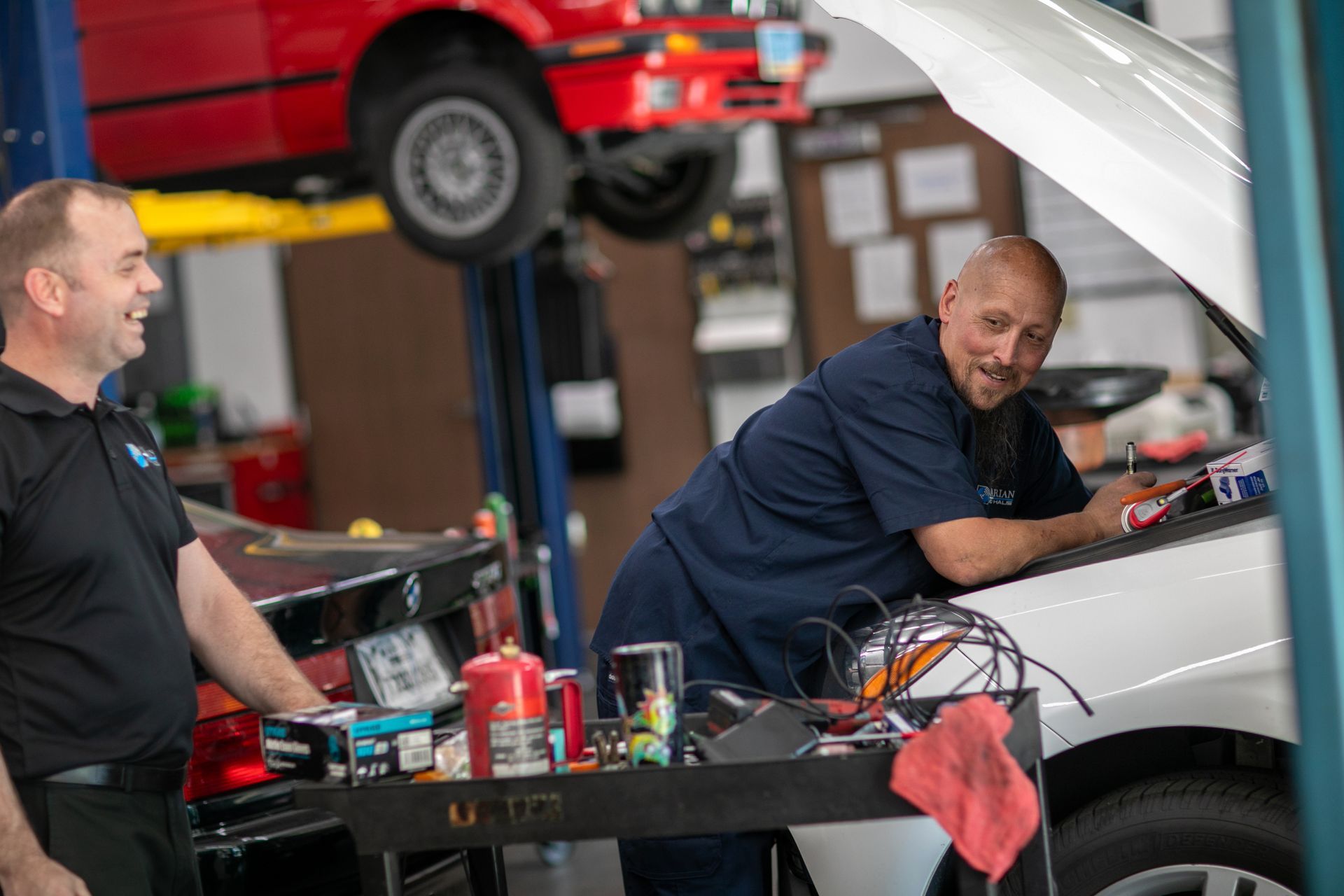 Two mechanics smiling while working on a white car in a garage with another vehicle lifted on a hydraulic lift overhead. | Bavarian Auto Haus