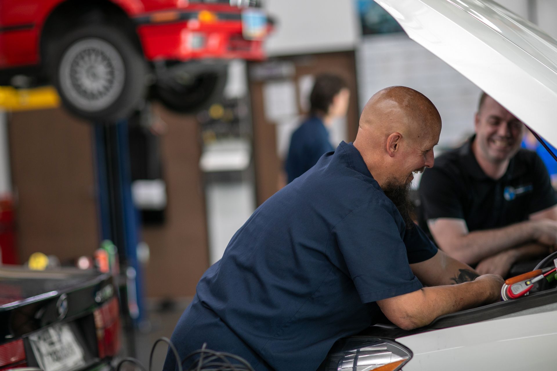 Two mechanics in a garage laugh while working on a car with its hood up, with another vehicle lifted in the background. | Bavarian Auto Haus