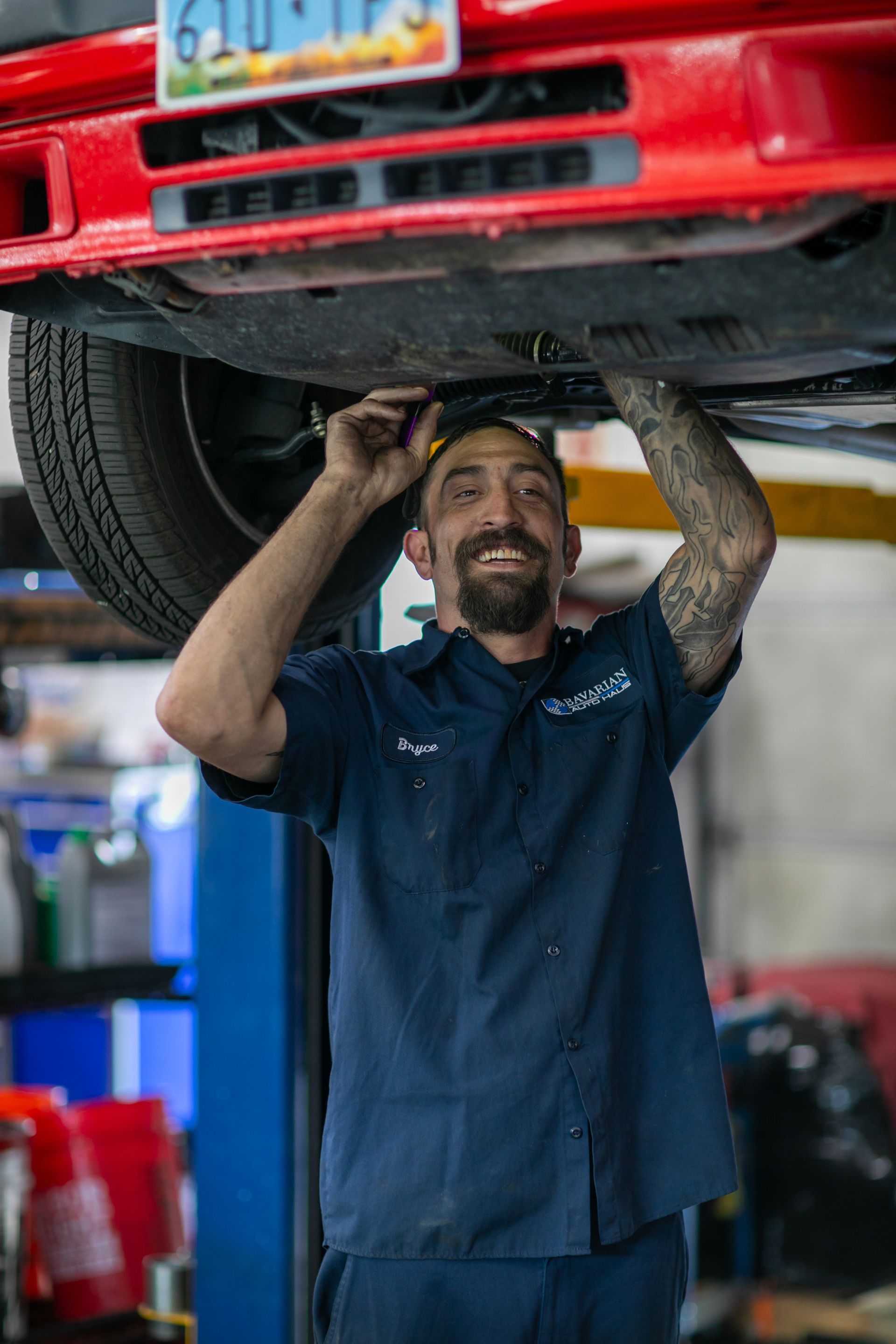 A smiling mechanic in a navy shirt repairs the underside of a red car lifted on a hydraulic rack in a garage. | Bavarian Auto Haus