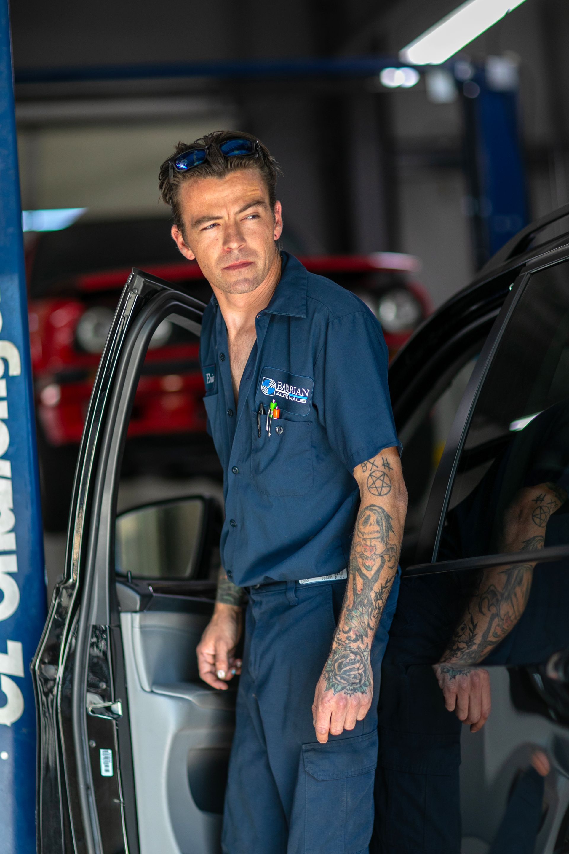 A tattooed mechanic in a dark blue uniform standing by an open car door in a garage, looking toward the side. | Bavarian Auto Haus