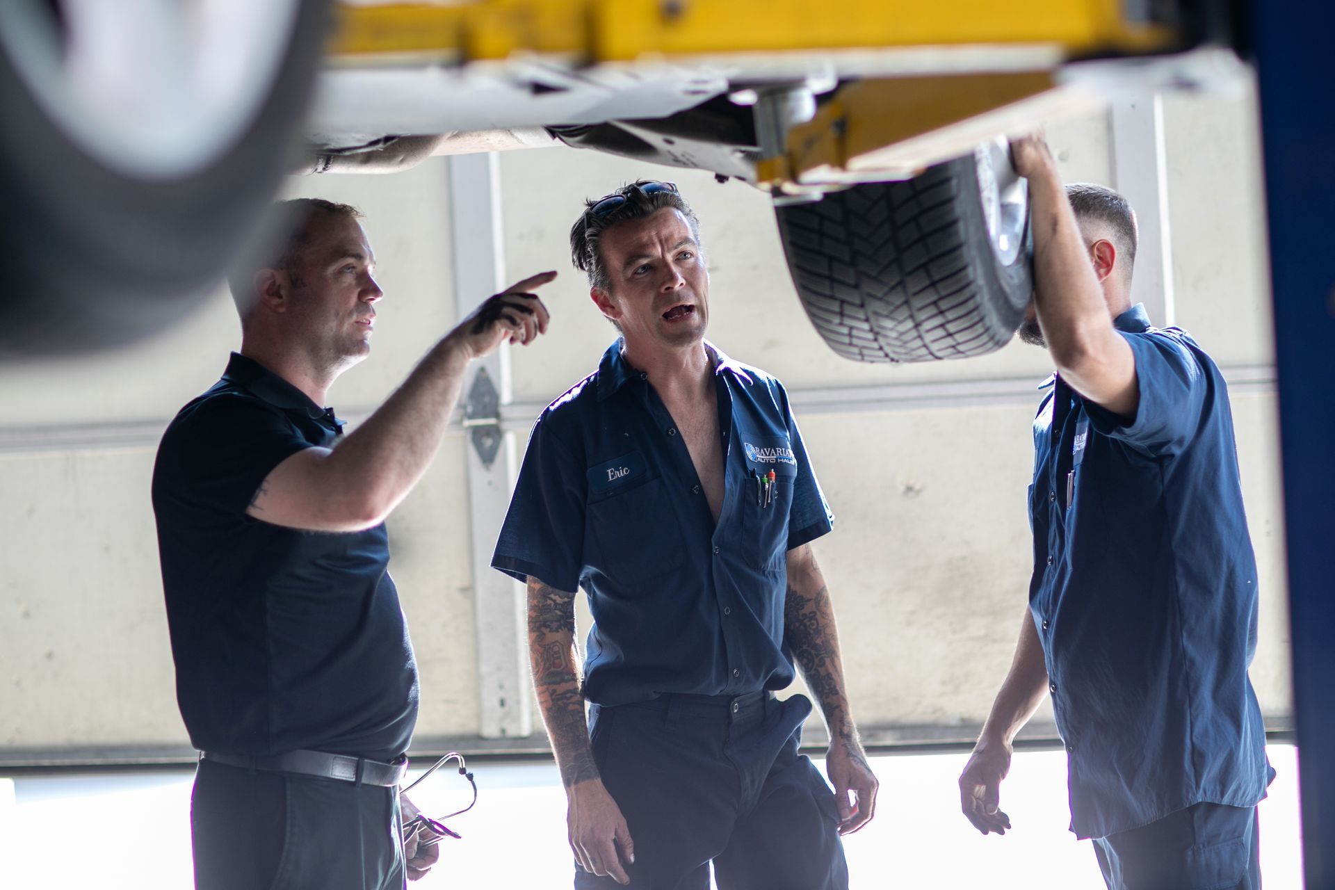 Three mechanics in uniforms stand in a garage, discussing repairs while pointing at the lifted vehicle's underside. | Bavarian Auto Haus