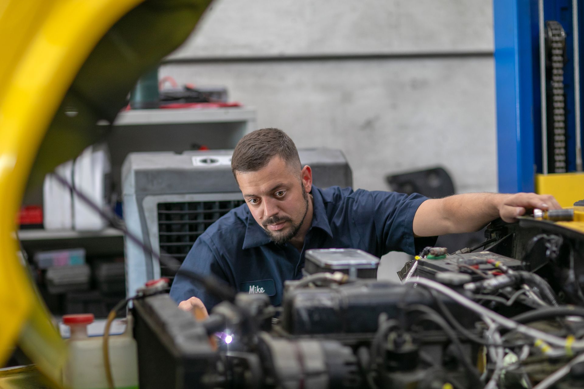 A focused mechanic in a blue uniform inspecting the open engine bay of a vehicle in an auto repair shop. | Bavarian Auto Haus