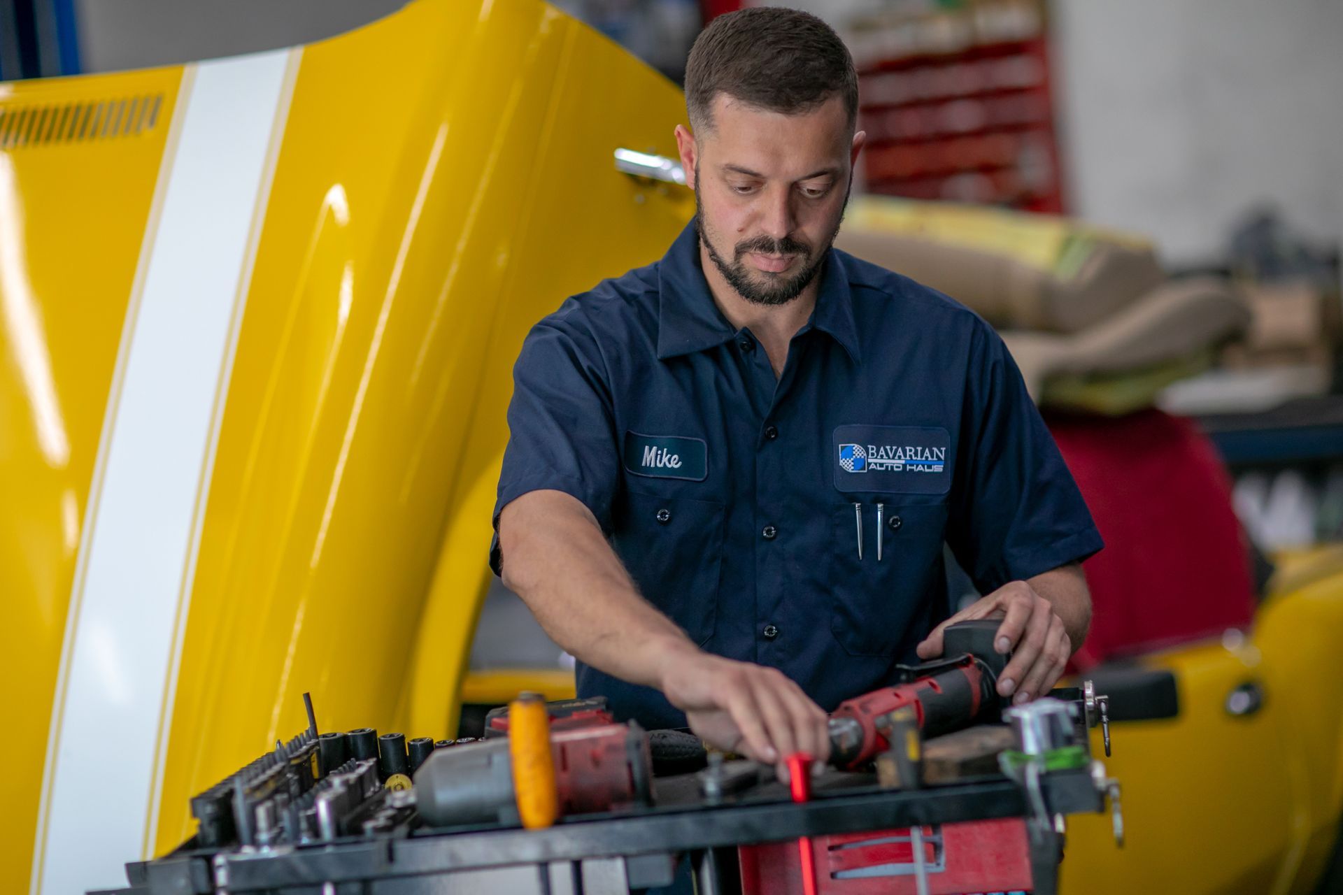 A mechanic in a blue uniform selects a tool from a rolling cart in a garage with a yellow car hood visible in the background. | Bavarian Auto Haus