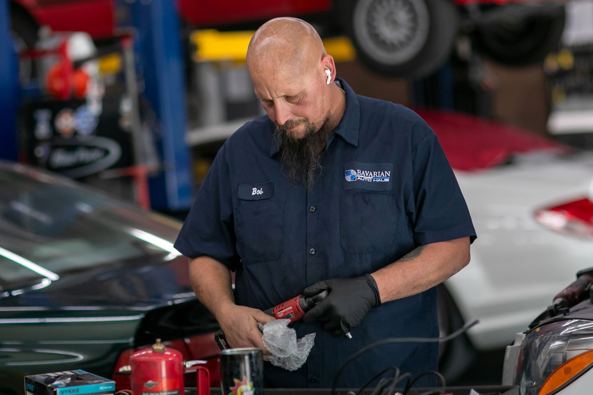 A mechanic in a blue uniform and black gloves works on a car part inside a workshop. | Bavarian Auto Haus