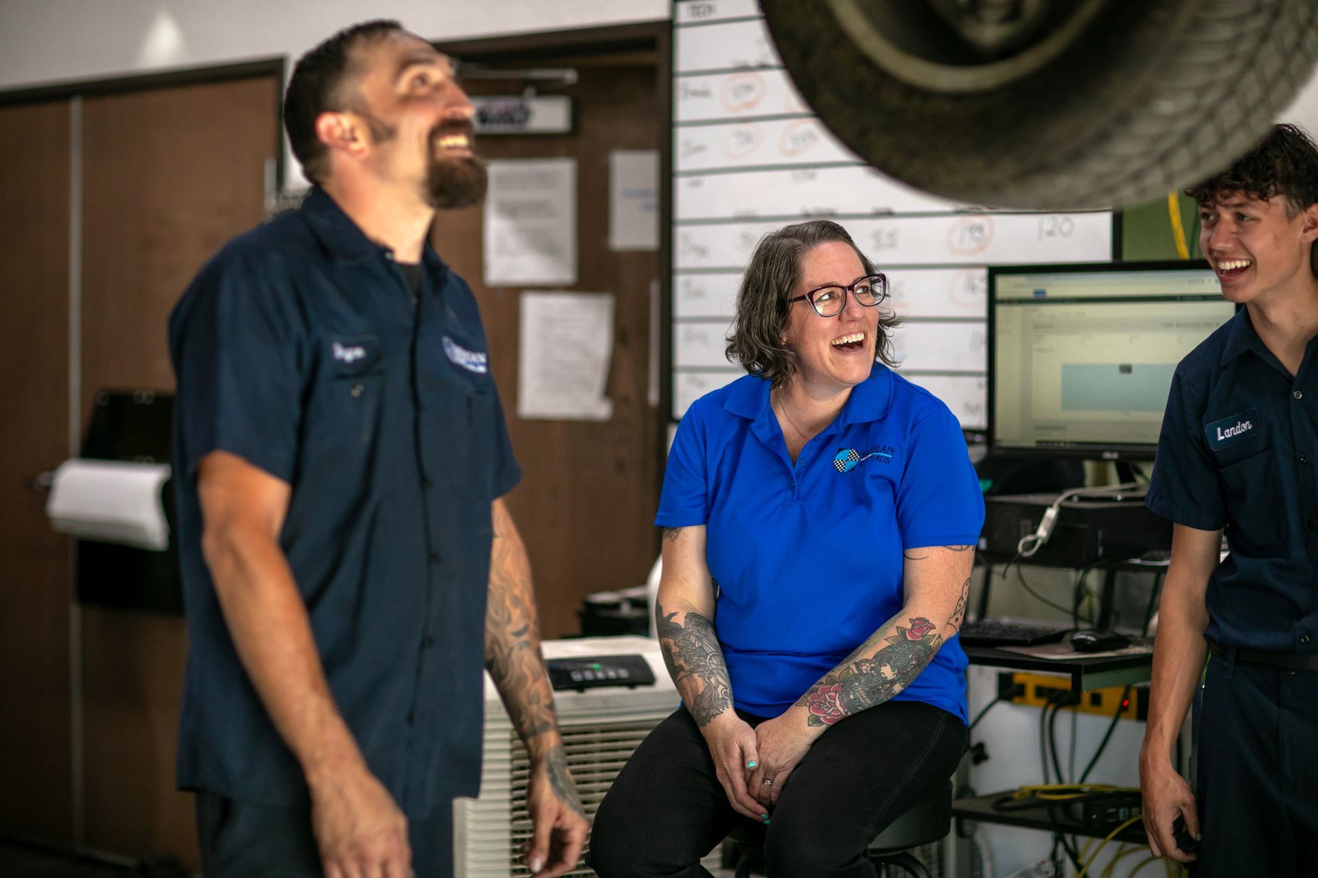 Three mechanics in a garage, laughing and looking upward at a car lifted on a hoist. | Bavarian Auto Haus