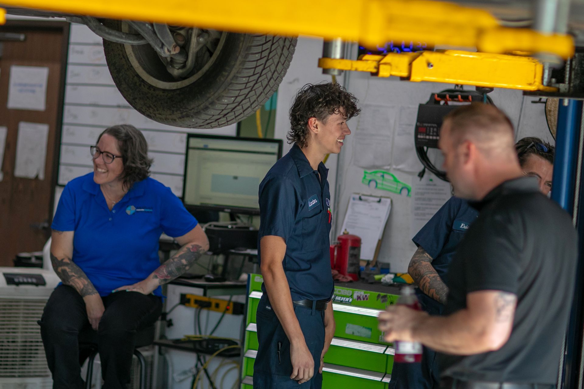 Three people in an auto shop consult while a car is lifted on a hydraulic rack above them. | Bavarian Auto Haus