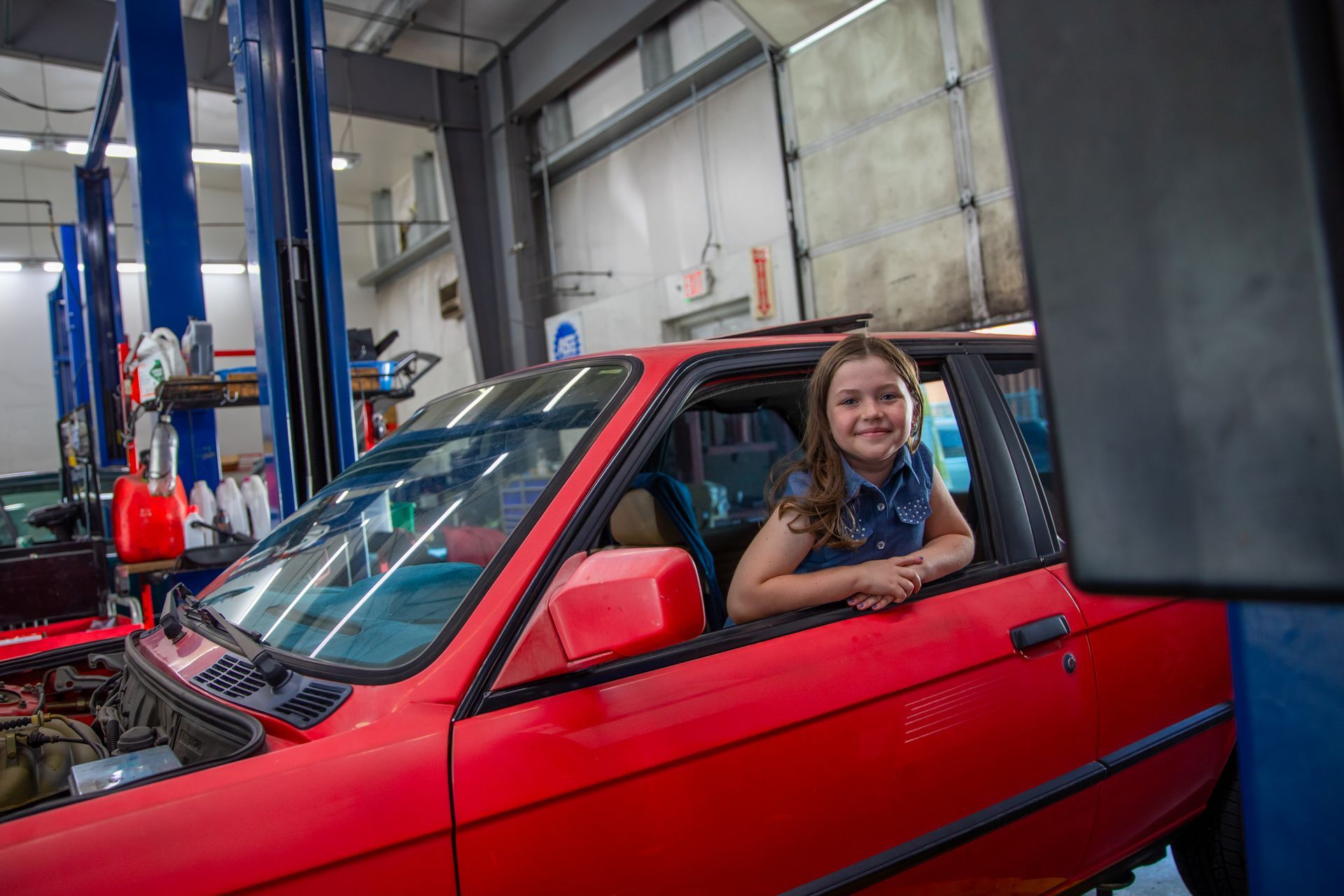 A smiling young person leans out the window of a bright red car in a mechanical garage with blue vehicle lifts. | Bavarian Auto Haus