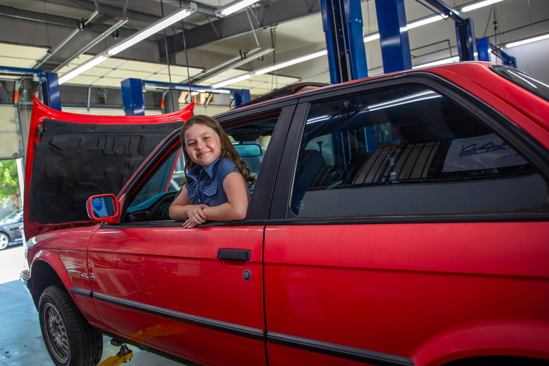 A smiling person sits in the driver's seat of a red car with its hood open inside an auto repair shop. | Bavarian Auto Haus