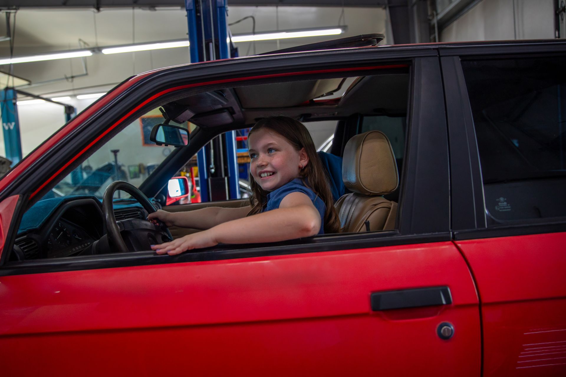 A smiling person sits in the driver's seat of a bright red car inside a repair shop. | Bavarian Auto Haus