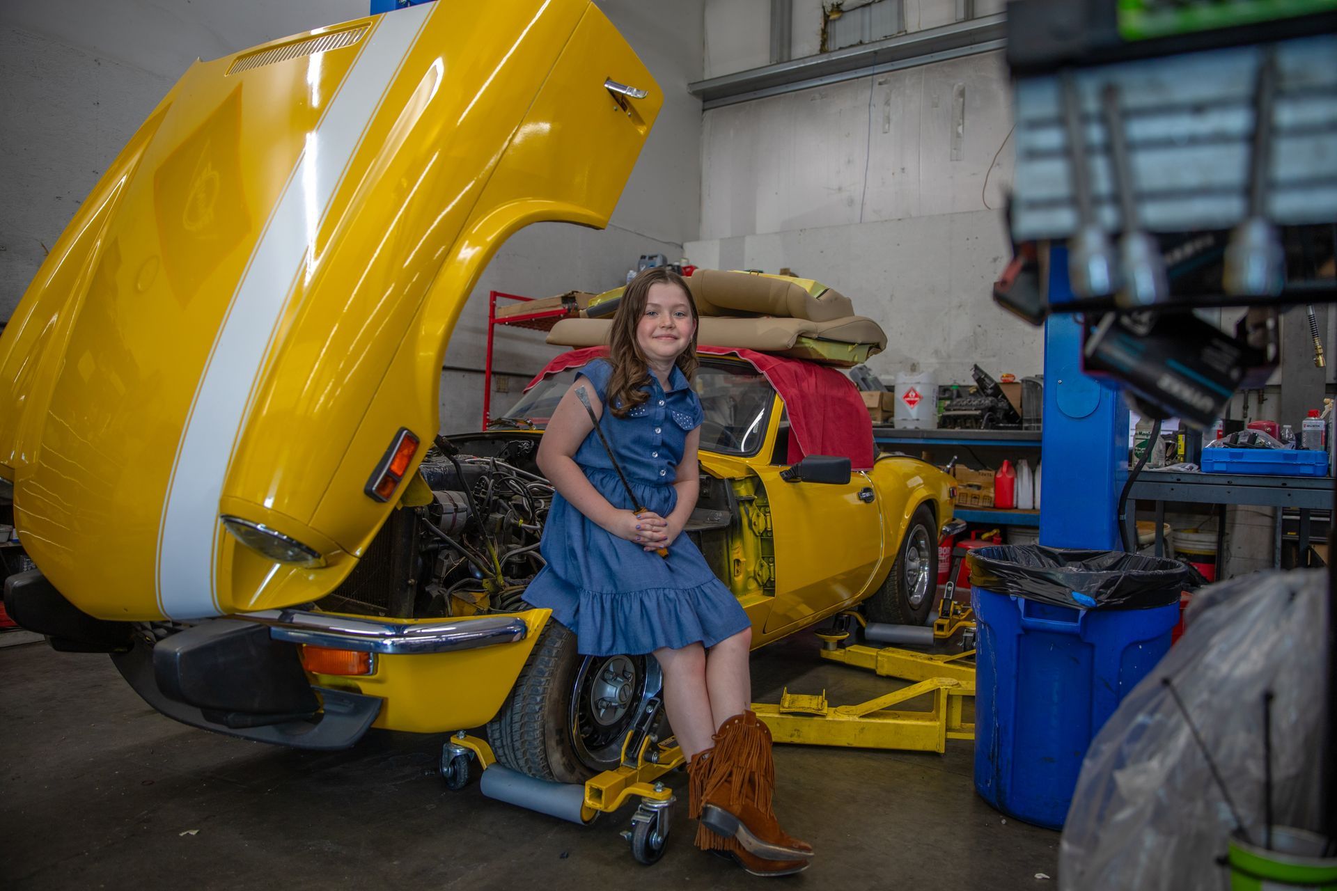 A smiling person in a blue dress and brown boots sits on a yellow car with its hood open inside a garage workshop. | Bavarian Auto Haus
