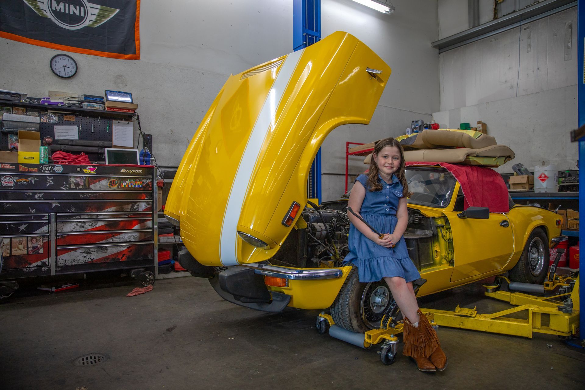 A smiling child in a blue dress and cowboy boots sits on the front tire of a yellow car with an open hood in a garage. | Bavarian Auto Haus