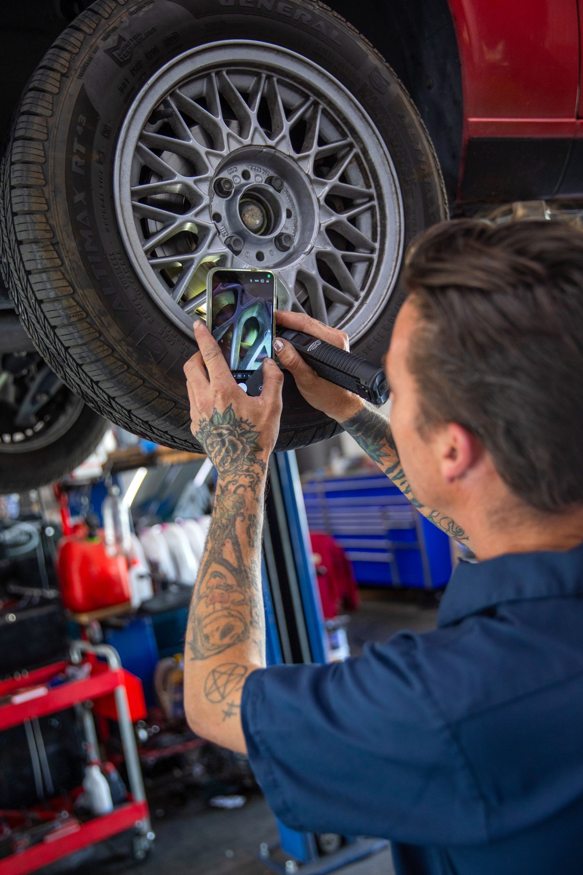 A mechanic with tattooed arms photographs the wheel of a car elevated on a lift in a workshop. | Bavarian Auto Haus