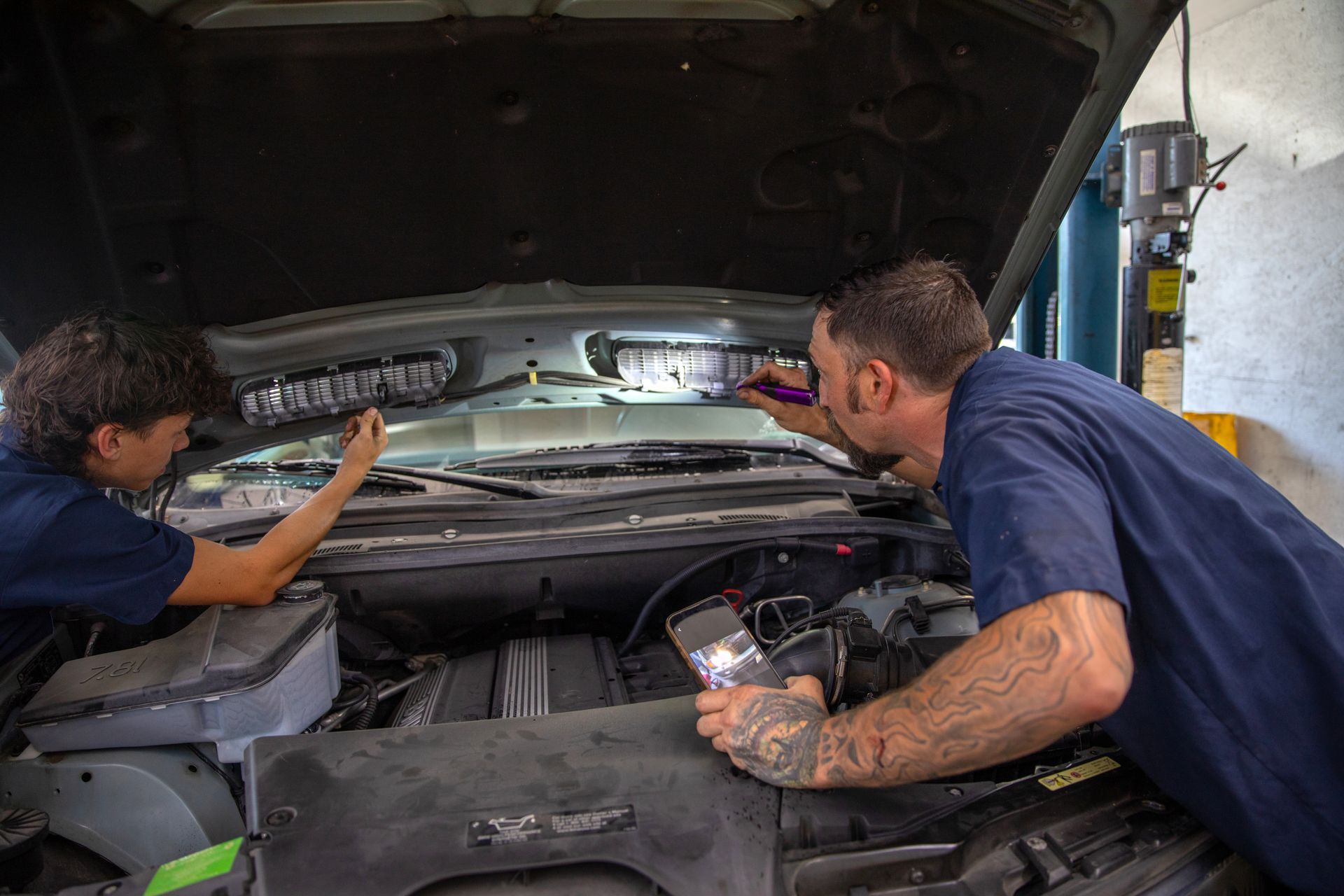 Two mechanics in dark blue shirts inspecting a car engine with flashlights in a repair shop. | Bavarian Auto Haus