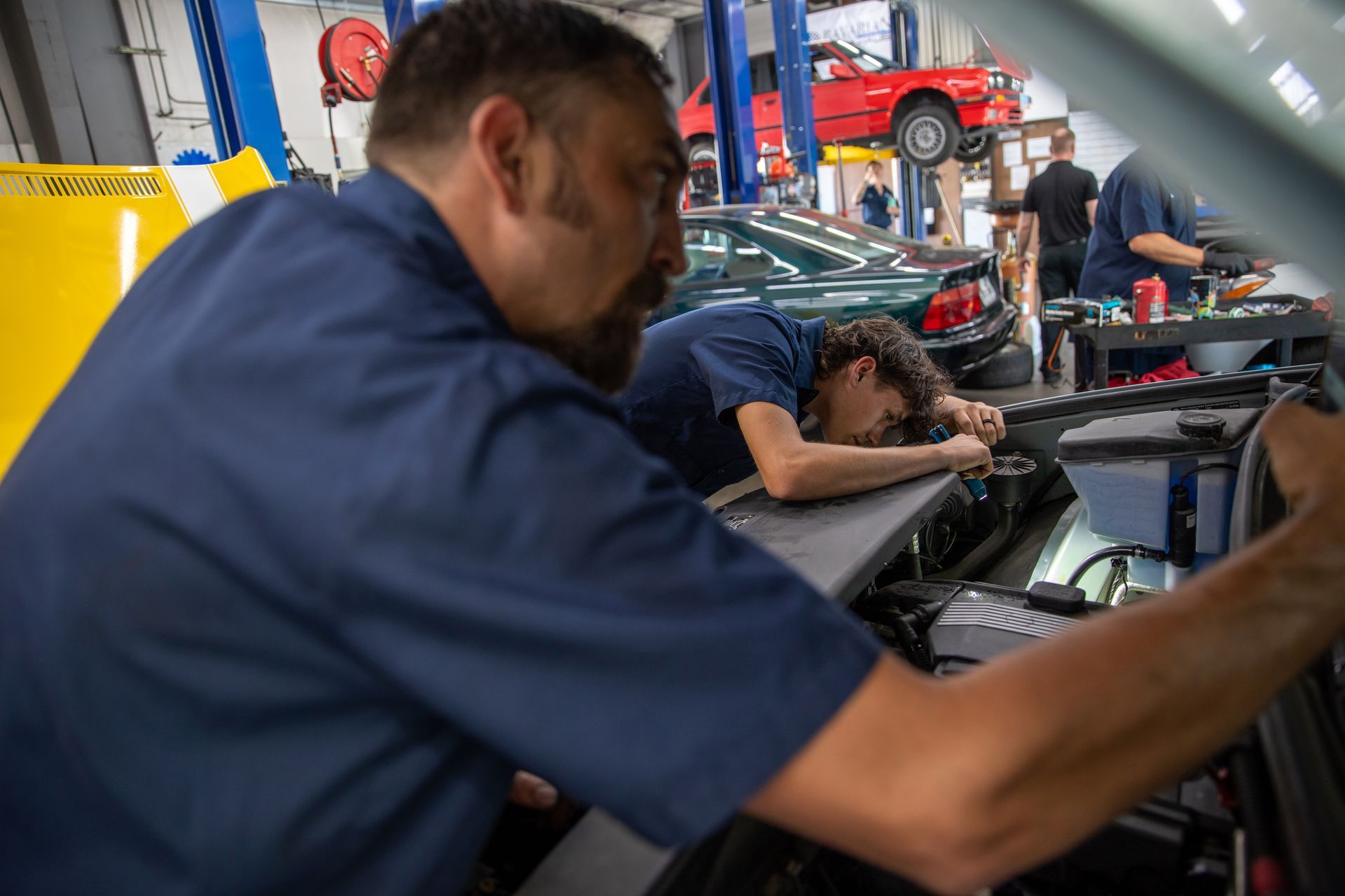 Two mechanics in a garage work under the open hood of a car, while other vehicles are visible on lifts in the background. | Bavarian Auto Haus