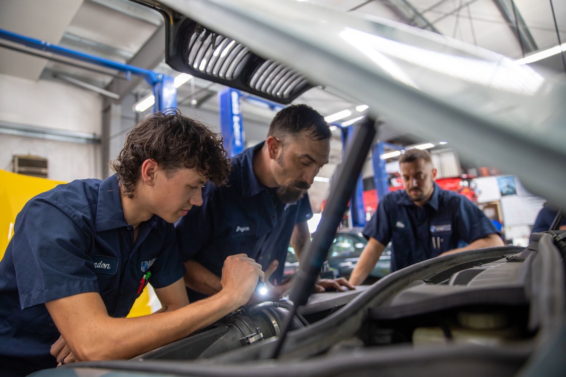 Three mechanics in blue uniforms inspect a car engine with a flashlight inside a bright auto repair shop. | Bavarian Auto Haus