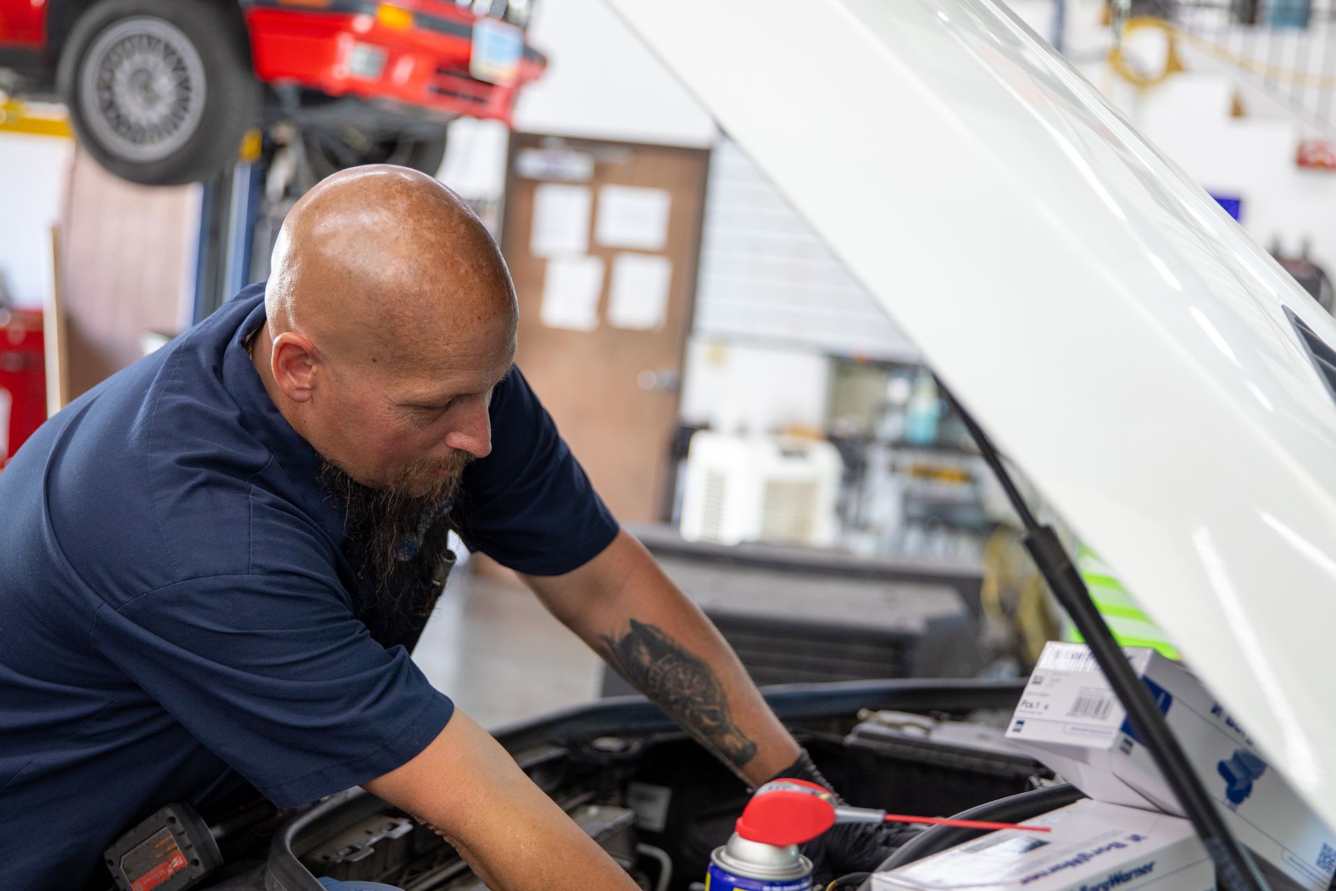A mechanic in a navy shirt works under the hood of a white vehicle in a brightly lit automotive repair shop. | Bavarian Auto Haus
