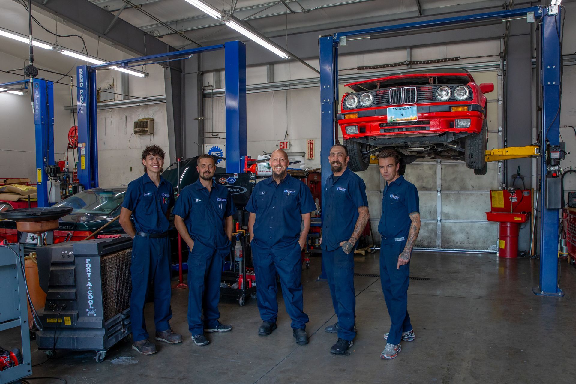 Five mechanics in blue uniforms stand in a row inside an auto repair shop with a red car lifted on a hydraulic rack. | Bavarian Auto Haus