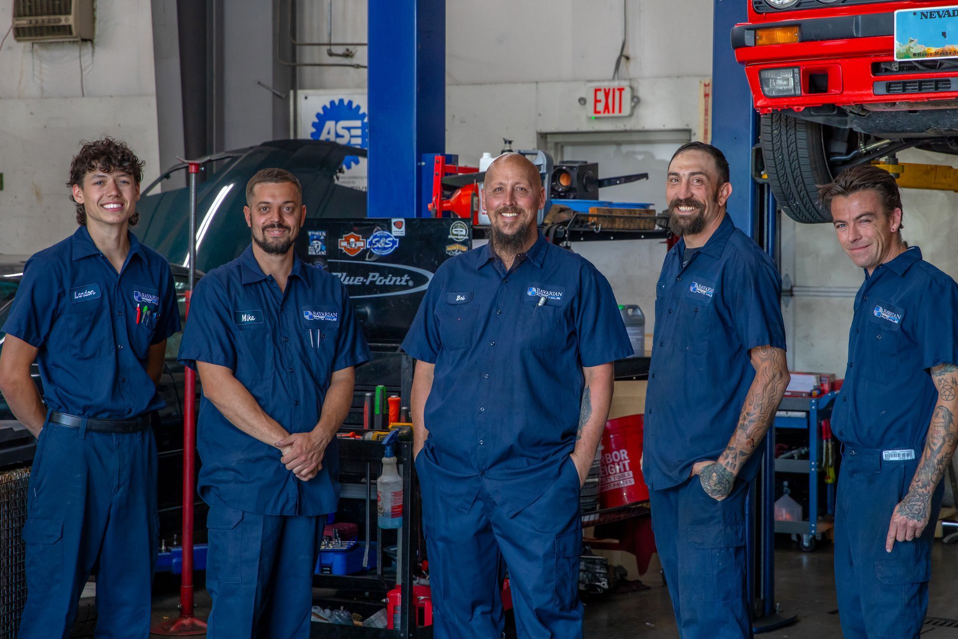 Five mechanics in matching blue uniforms stand together and smile in an auto repair shop with a car on a lift. | Bavarian Auto Haus