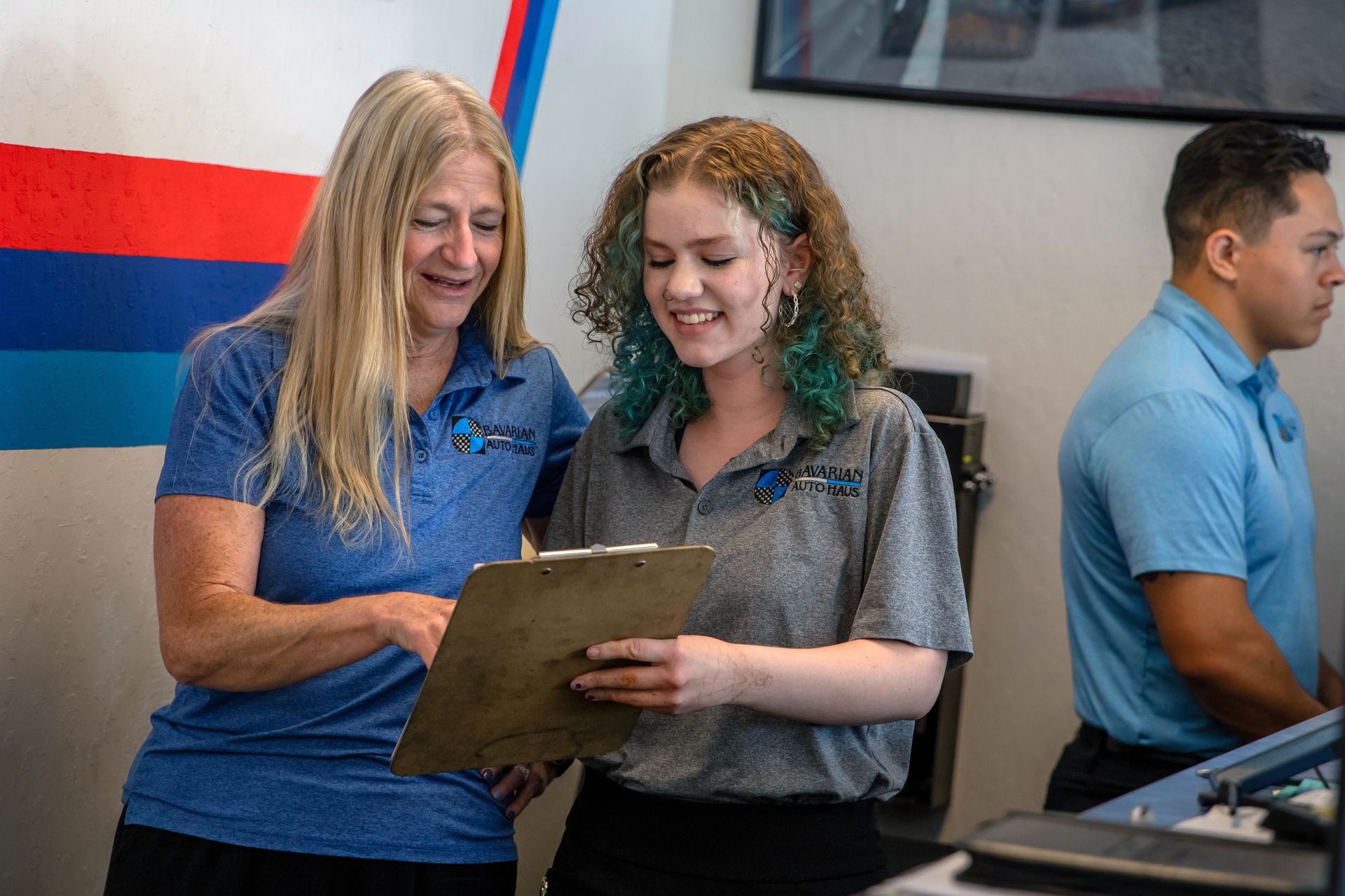 Two colleagues in uniform stand in an office, reviewing notes on a clipboard and smiling while a third person works nearby. | Bavarian Auto Haus