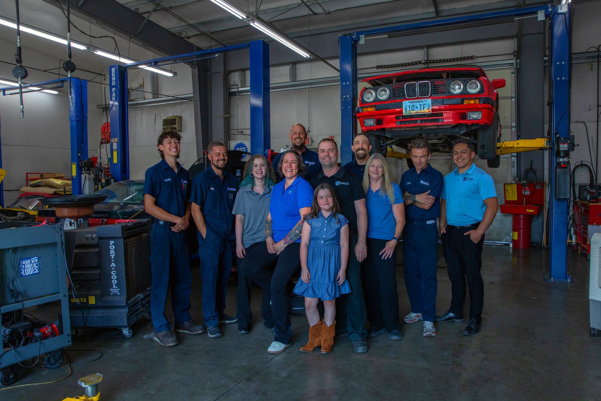 A group of eleven people standing together in an auto repair shop with a red vehicle lifted on a rack in the background. | Bavarian Auto Haus
