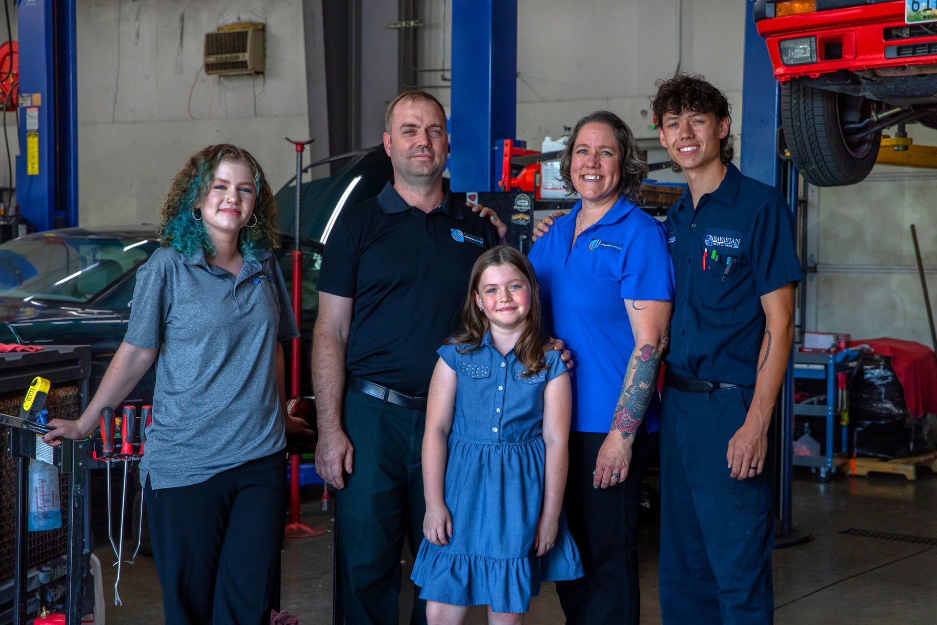 Family in auto repair shop, posing together. Blue and gray clothing. Car parts and tools visible. | Bavarian Auto Haus
