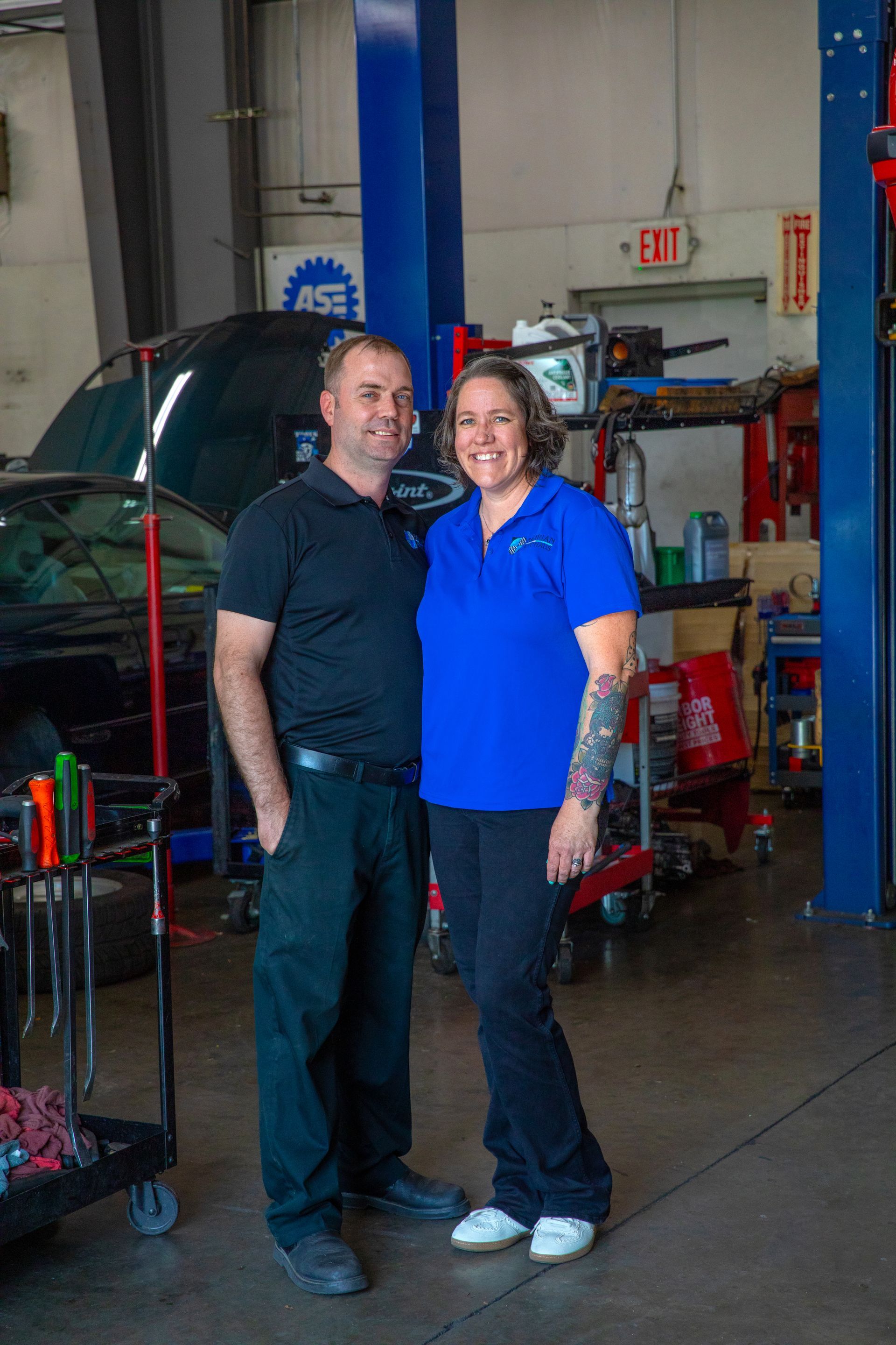 A smiling couple stands together in a car repair shop with a vehicle lift and tools in the background. | Bavarian Auto Haus