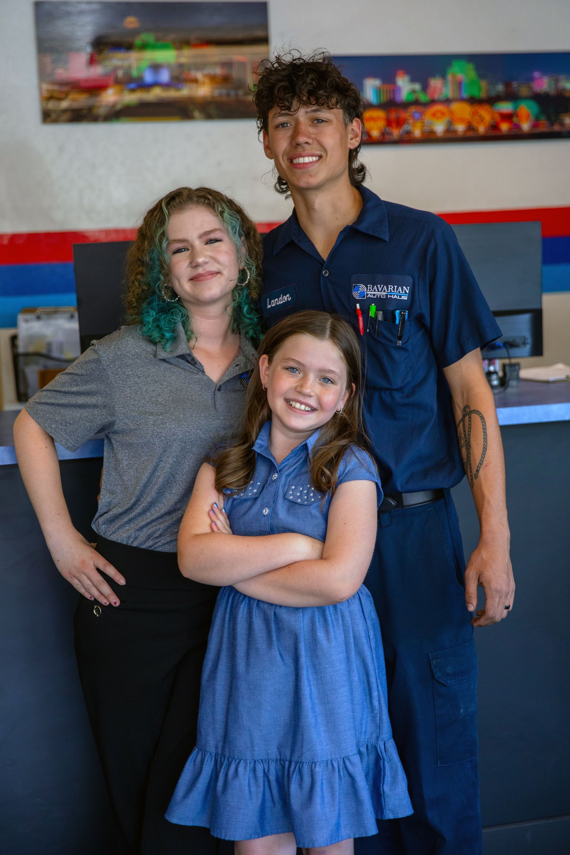 Three people standing together in an office setting; a young girl in the front smiles with arms crossed. | Bavarian Auto Haus