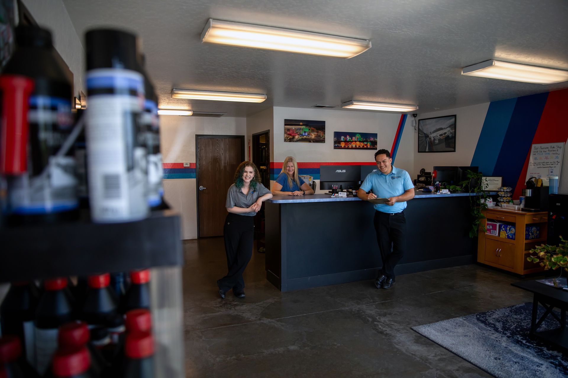 Three employees stand behind a service desk in an auto shop with blue and red wall accents and overhead fluorescent lights. | Bavarian Auto Haus
