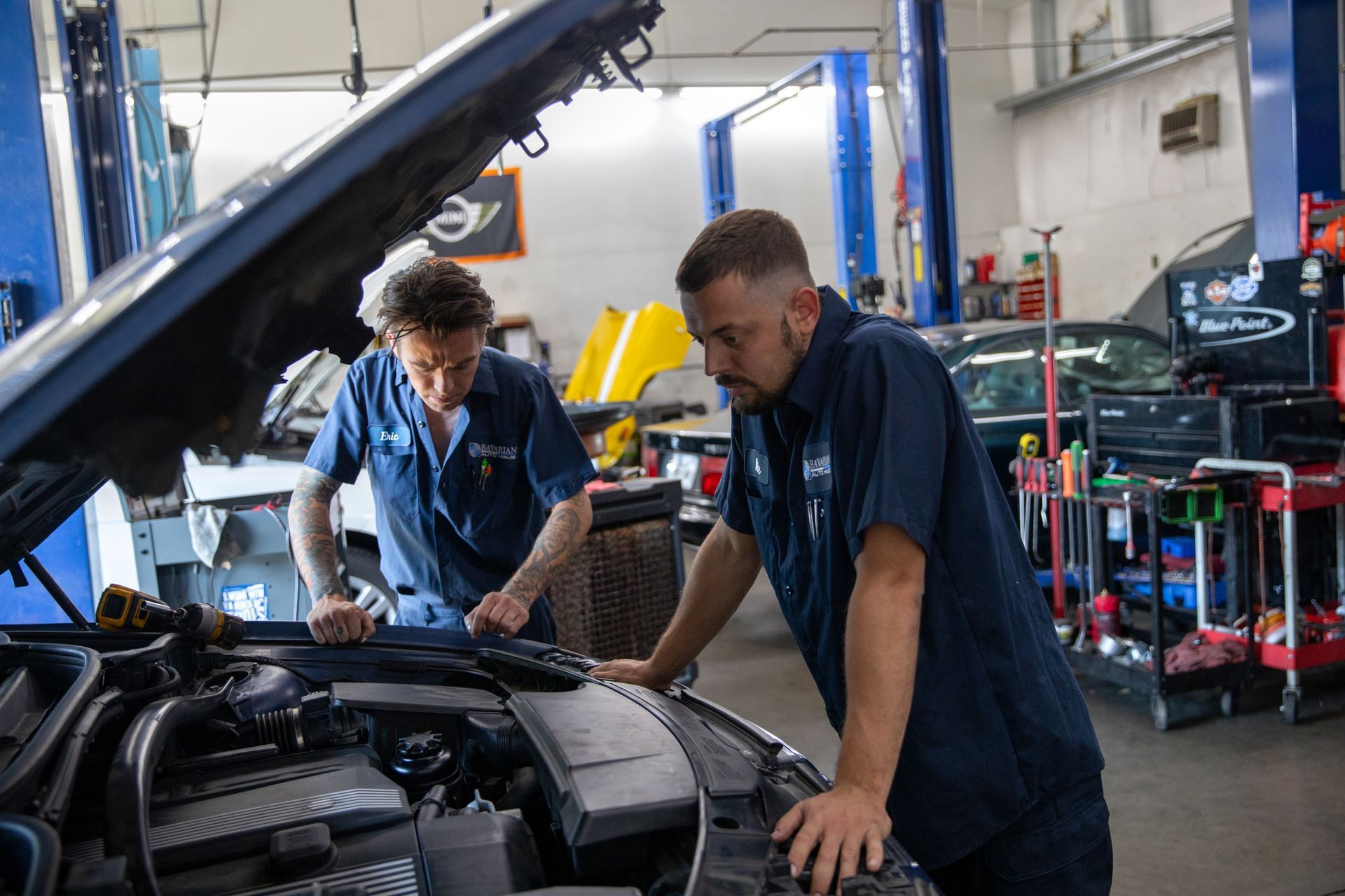 Two mechanics in blue uniforms stand at an open car hood in a repair shop, inspecting the engine together.  | Bavarian Auto Haus